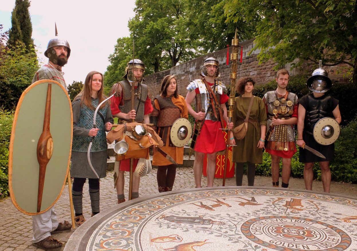 Eight people in Roman-style armour and tunics pose with shields, spears and equipment beside a circular mosaic and stone wall.
