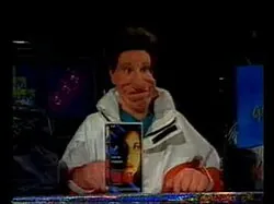 Vito sits behind a studio desk in a pale jacket, with a boxed game standing in front of him.