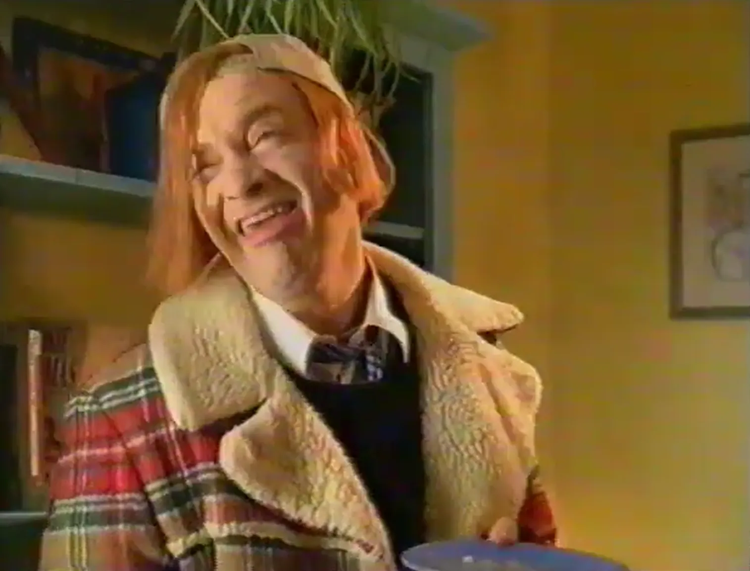 Close-up of a smiling young person with shoulder-length reddish hair and a backward beige cap, wearing a red plaid coat with a cream fleece collar over a shirt, tie and dark sweater in a warmly lit room.