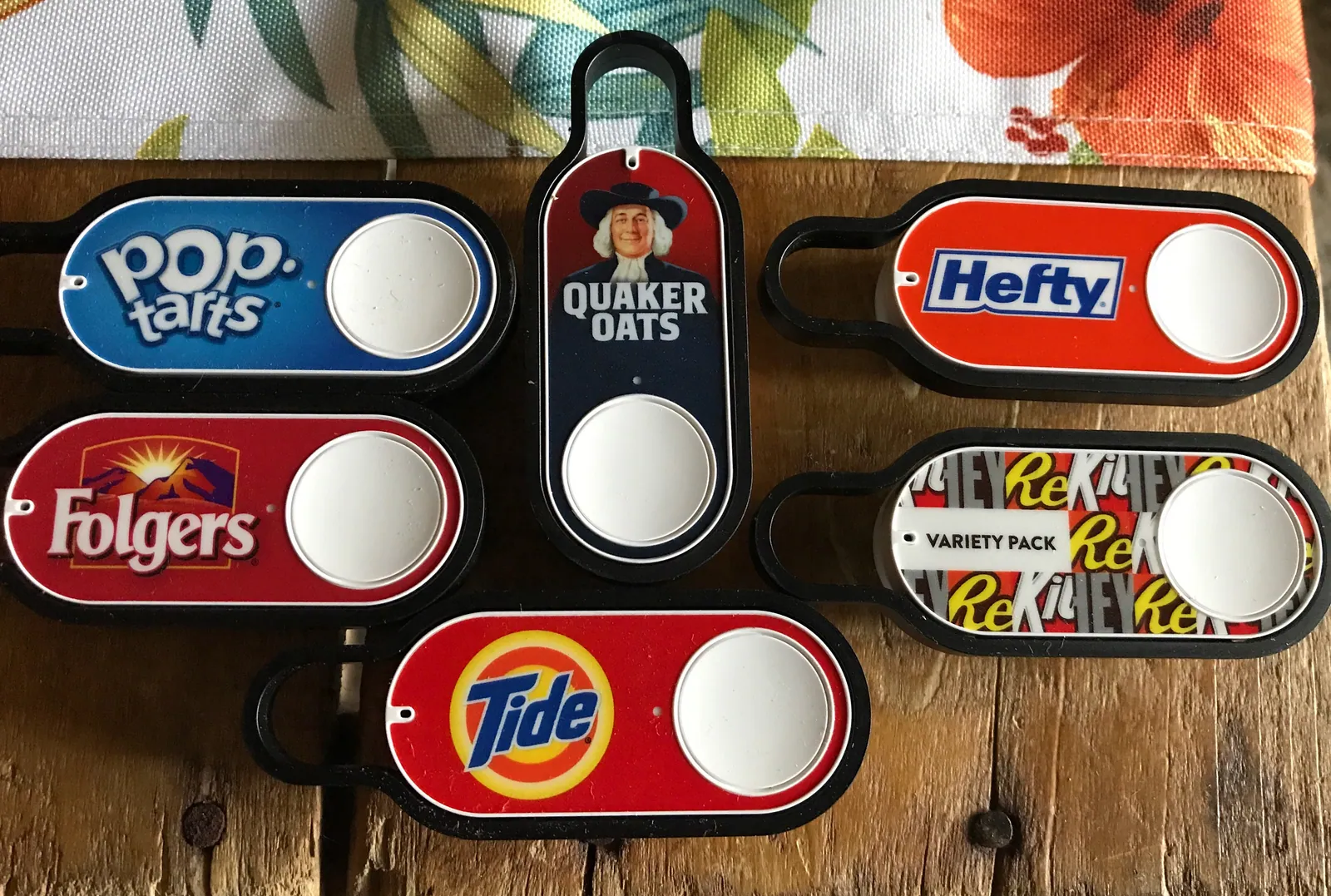 Several overlapping Amazon Dash Buttons lie on a wooden surface, including visible Pop-Tarts, Quaker Oats, Hefty, Folgers, variety pack, and Tide designs with white circular buttons.