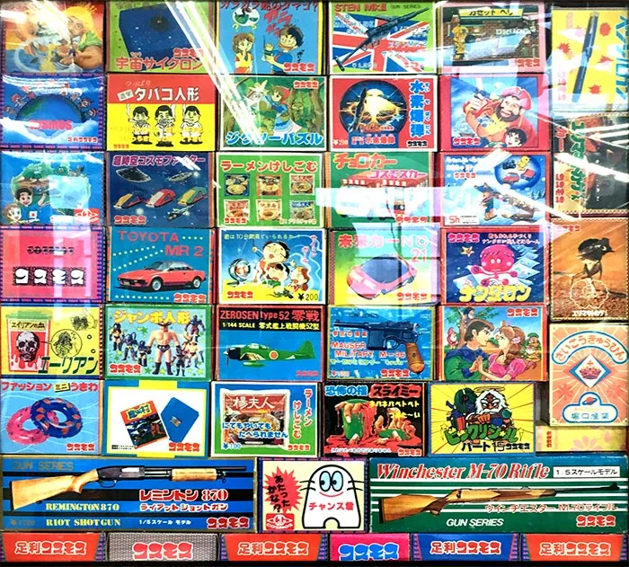 Grid of colorful Japanese toy package fronts or display cards behind a reflective surface. Repeated red Cosmos logos appear across illustrations of vehicles, figures, weapons, puzzles and novelty items, with several 100-yen and 200-yen markings.