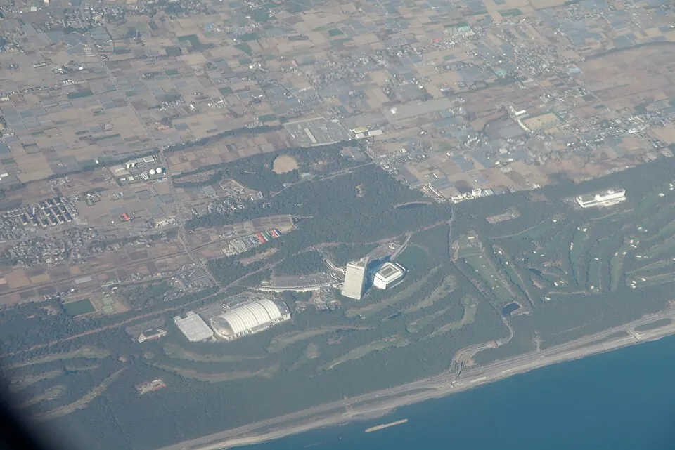 Aerial view of a coastal resort, with a long white-roofed building, hotel tower, forest belt and beach.