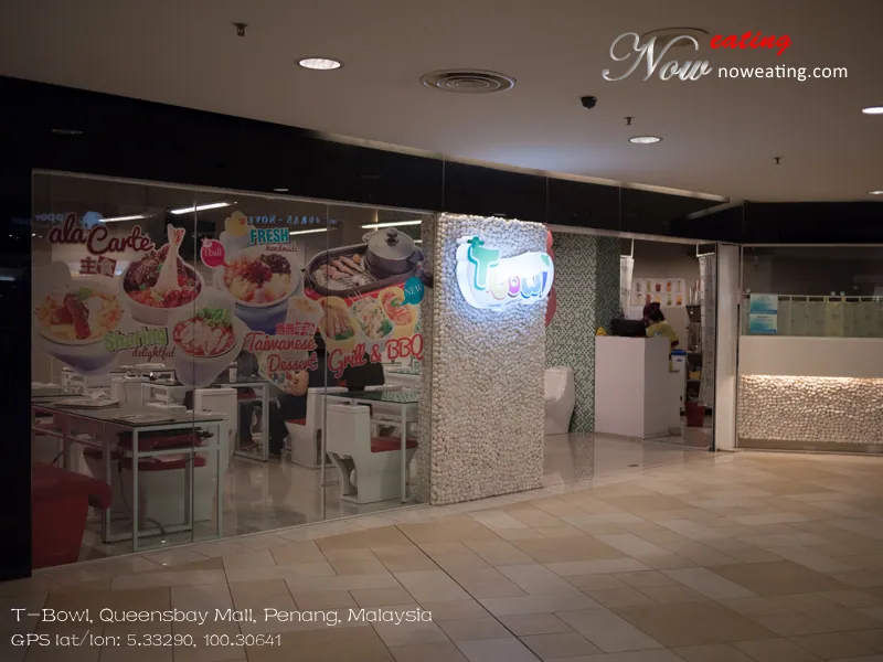 Mall-facing restaurant entrance with an illuminated T-Bowl logo, illustrated window panels, white toilet-shaped seats, glass tables and a reception counter. A Now Eating watermark appears at upper right, with Queensbay Mall location text and coordinates along the bottom.