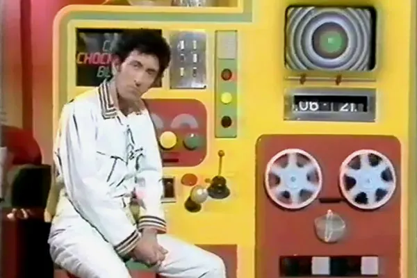 A man in a white jumpsuit sits beside a large yellow and red computer-like control panel with a circular screen, rows of buttons, levers, and two large reel-shaped wheels.