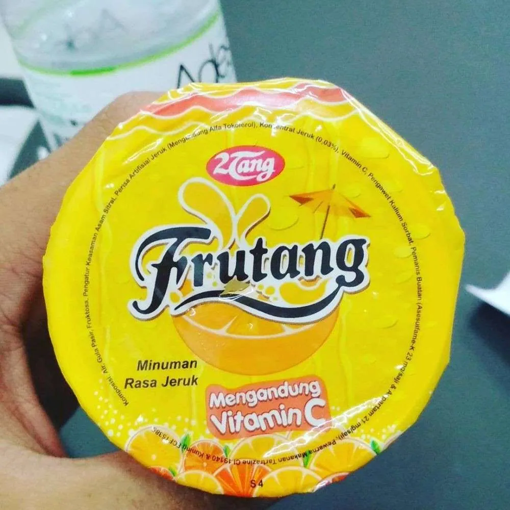 Close-up of a yellow Frutang orange drink foil lid held in a hand, with the 2Tang logo, orange illustrations and vitamin C branding.