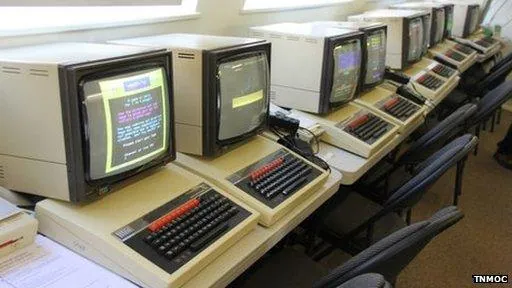A row of beige BBC Microcomputer workstations with CRT monitors, keyboards with red function keys, and black chairs arranged along a table beside windows.