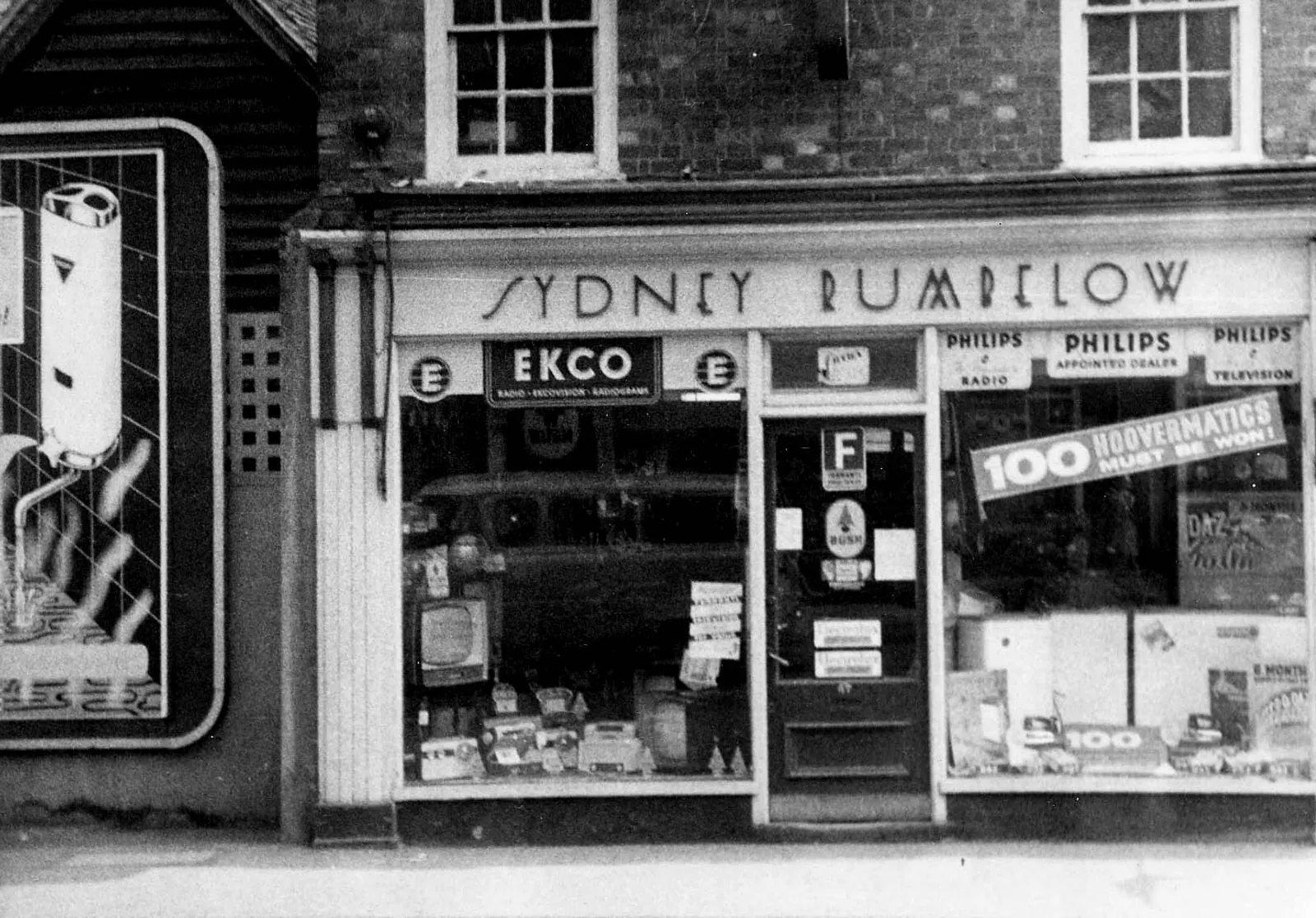 Black-and-white photograph of a shop with a SYDNEY RUMBELOW sign above the entrance, Ekco and Philips signs in the windows, and displays including a television, radios and large household appliances.