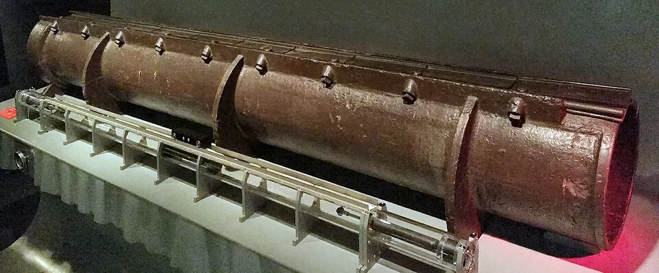 Museum display showing a long rust-brown cylindrical atmospheric railway pipe behind a cutaway mechanical rail assembly.