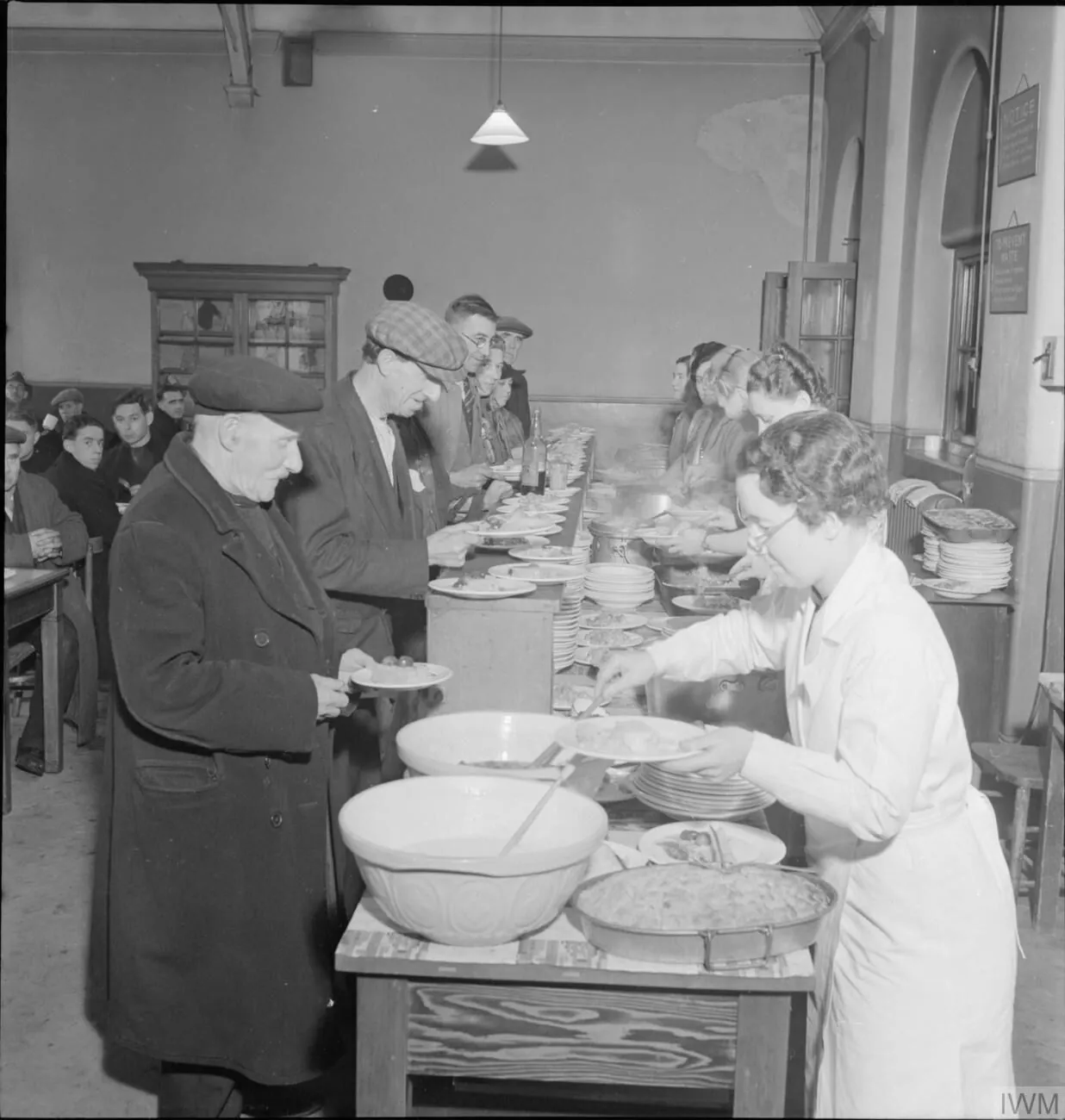 An older man holds a plate at a long serving counter while staff dish out food and other diners queue.