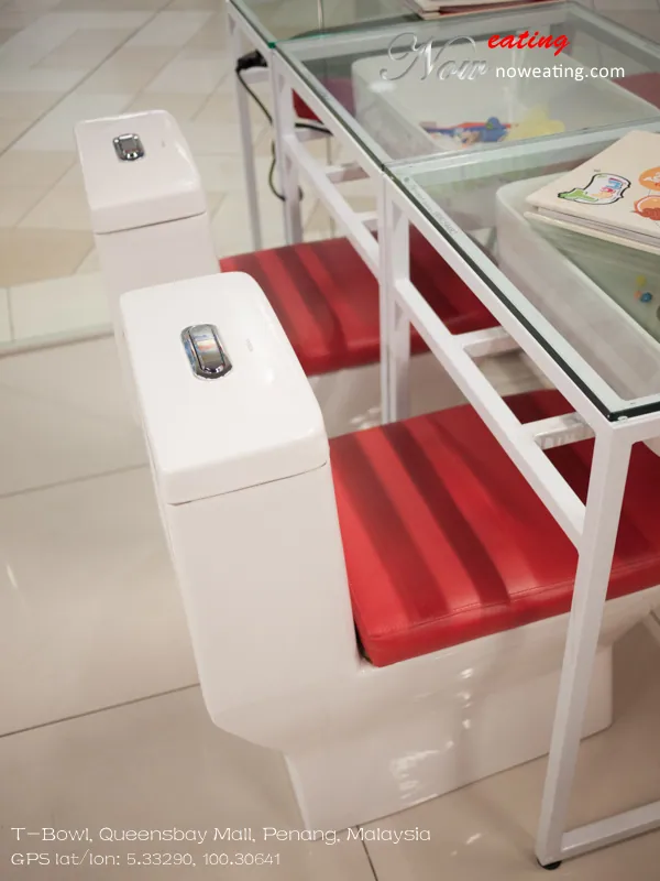 Close view of two glossy white toilet-shaped seats with rectangular tanks and red padded cushions beside a white-framed glass table. Part of an illustrated menu lies on the tabletop. A Now Eating watermark and Queensbay Mall location overlay are visible.