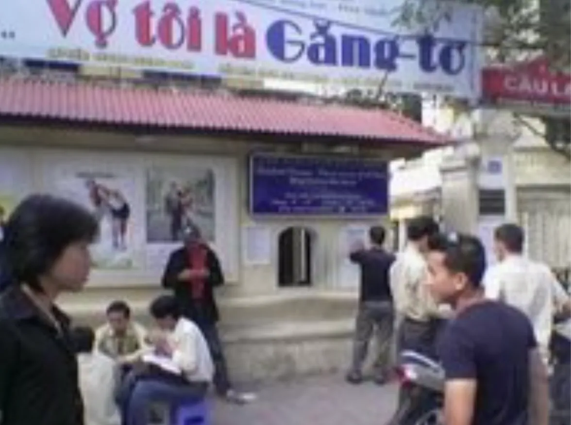 People standing and sitting outside the Fansland Cinema ticket office in Hanoi, beneath a red-tiled roof and Vietnamese-language signage.