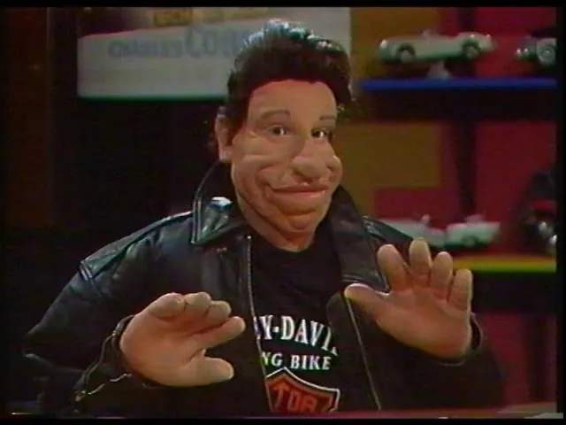 Vito, a puppet with slick dark hair and exaggerated facial features, gestures in a black leather jacket in the Game Over studio.