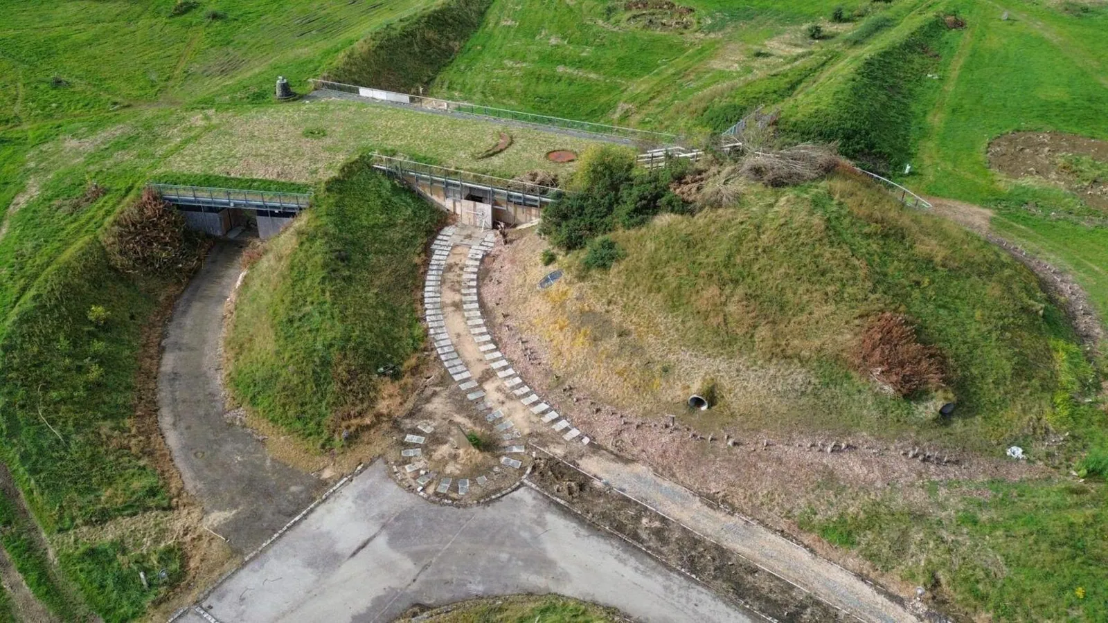 Aerial view of grass-covered mounds with two recessed entrances, metal railings, a curving stepped path and surrounding paved and grassy ground.