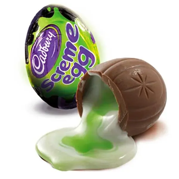 Foil-wrapped Cadbury Screme Egg beside a broken milk-chocolate egg with white fondant and bright green filling spilling onto a white background.