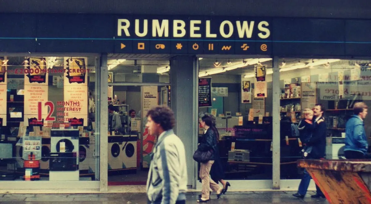 Colour photograph of a Rumbelows high-street shopfront with yellow lettering, appliance-filled windows, large credit and interest-free posters, and several pedestrians passing outside.