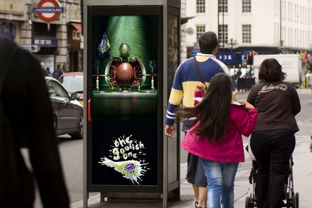 Pedestrians pass a street advertising panel showing a large brown egg restrained in a chair under green light, with Cadbury Screme Egg branding near the bottom; cars and an Underground sign appear behind them.