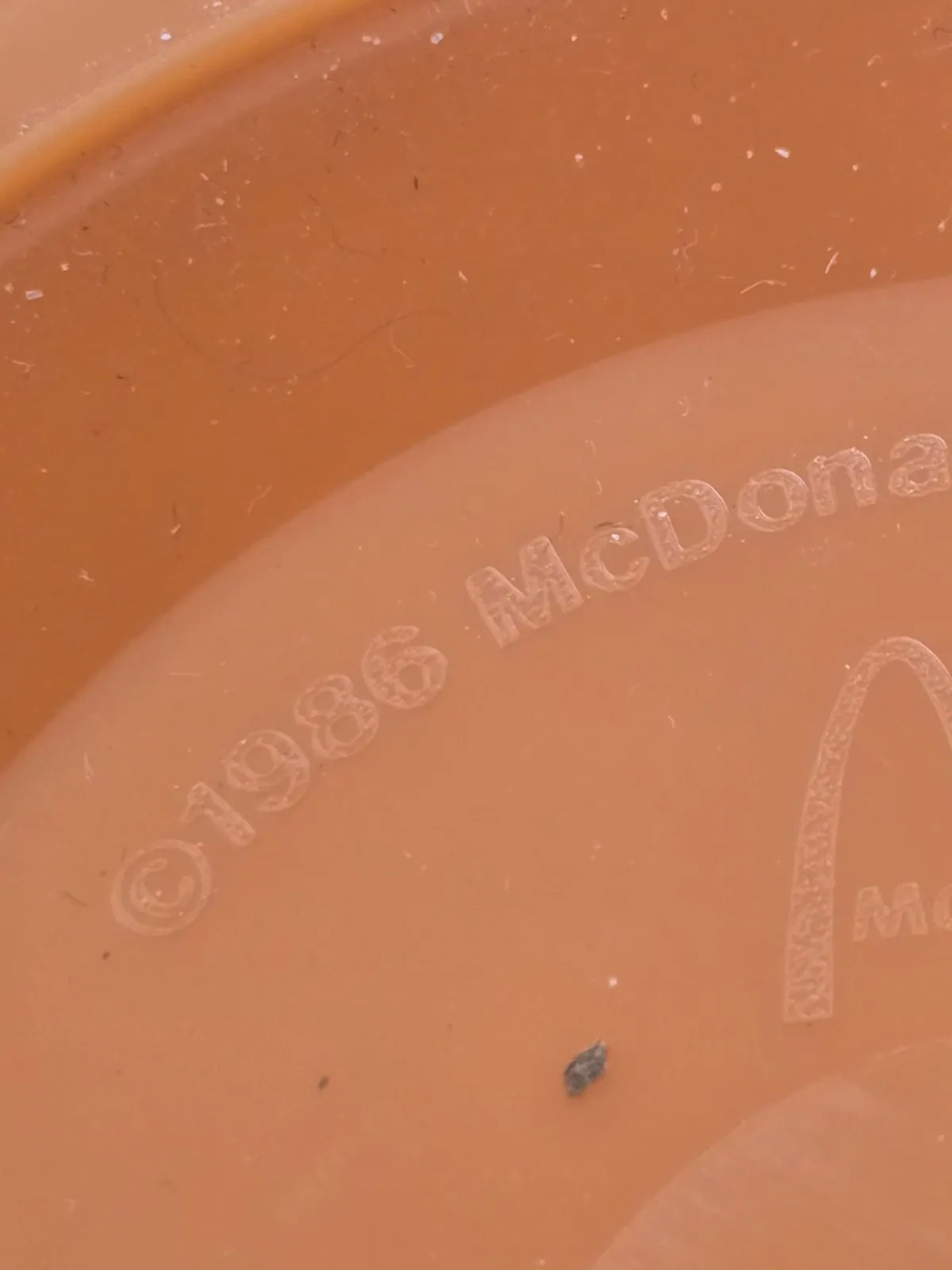 Extreme close-up of scratched and dusty orange plastic embossed with “©1986 McDonalds,” beside part of an embossed arch-shaped logo.