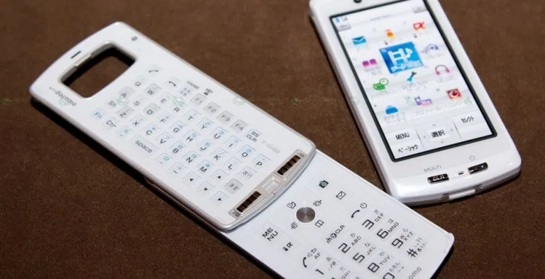 White modular mobile phone on a brown surface, with the touchscreen display detached beside a keyboard section opened to show QWERTY and numeric keypads.