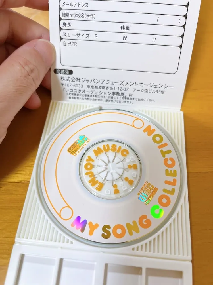 Open white plastic CD case held in one hand, containing a small disc with orange lettering and a printed recording-information card.