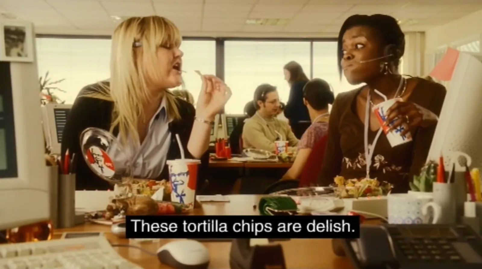 Two women wearing telephone headsets sit at desks in a busy office, eating from KFC containers. Computers, telephones and other workers fill the background, and a subtitle reads, “These tortilla chips are delish.”