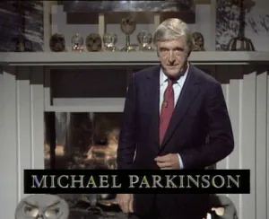 Michael Parkinson stands in a dark suit and red tie before a lit fireplace decorated with skulls; a black caption bar reads “Michael Parkinson.”
