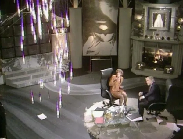 High-angle view of a television studio where a woman and a grey-haired man sit in office chairs beside a glass table, while bright vertical streaks obscure the left side of the picture and papers lie across the floor.