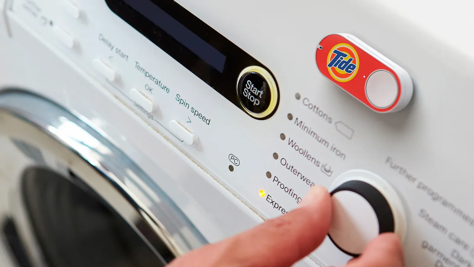 A Tide-branded Amazon Dash Button attached to a white washing machine above its controls, with a hand turning the programme dial.