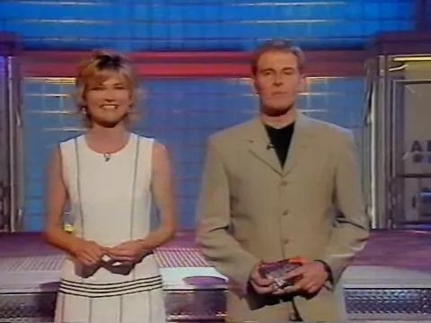 A smiling woman in a sleeveless white outfit and a man in a beige suit holding a small card stand on a blue-and-red television studio set.