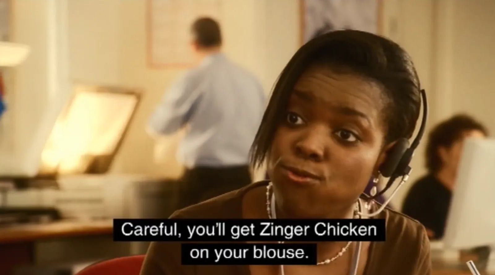 Close-up of a woman wearing a telephone headset in an office, with blurred workers and computer monitors behind her. A black subtitle reads, “Careful, you’ll get Zinger Chicken on your blouse.”