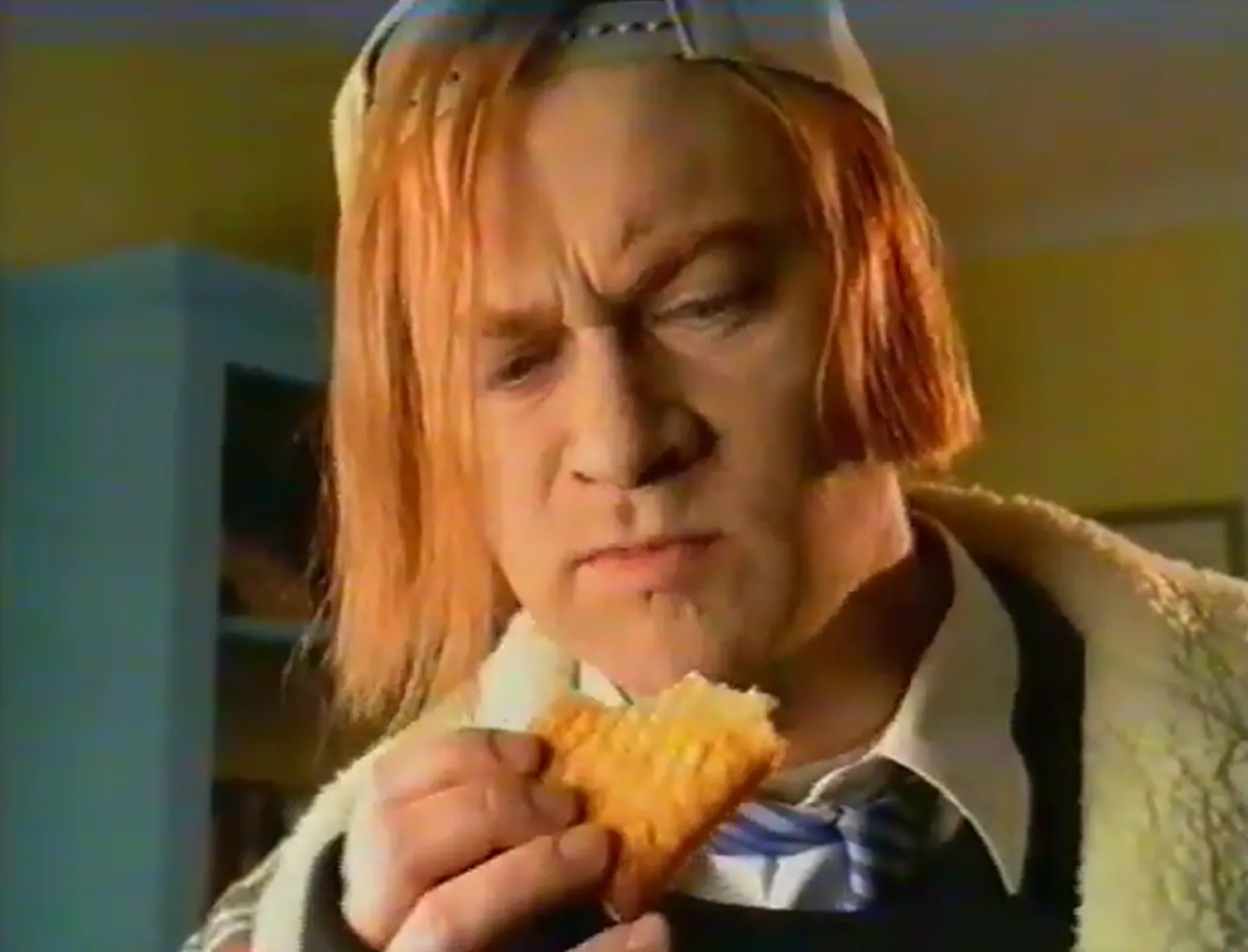 Close-up of a person with chin-length orange hair, sunglasses on top of their head, a striped tie and a fleece-lined jacket, examining a small rectangular toasted pastry held near their mouth.