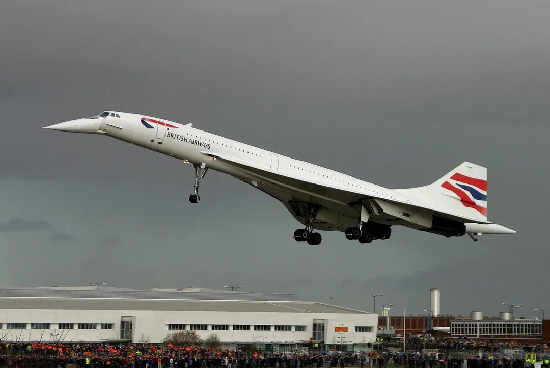 British Airways Concorde flying with its landing gear extended above airport buildings and a large crowd under a grey sky.