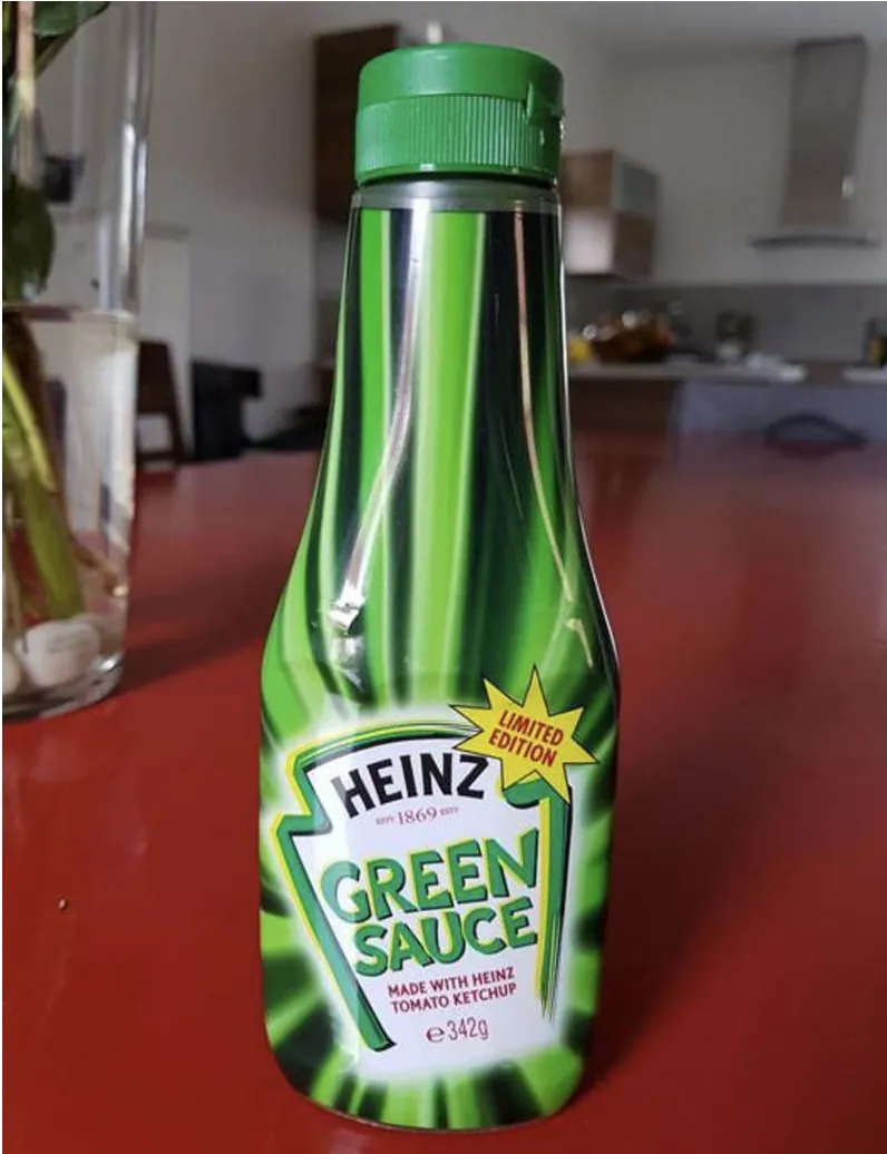Front view of a green-capped Heinz squeeze bottle on a red tabletop. Its green burst-pattern label reads “Heinz Green Sauce,” “Limited Edition,” “Made with Heinz Tomato Ketchup,” and “342g,” with a kitchen blurred in the background.