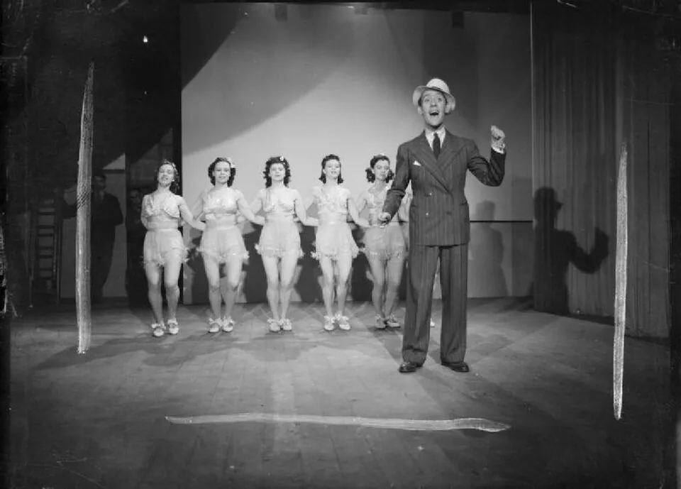 Tommy Trinder performs on a small stage with six women lined up behind him.