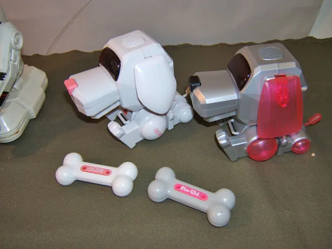 Two angular plastic robot dogs sit facing one another on dark fabric, one white with pink accents and one silver with translucent red ears and joints. Two white bone-shaped accessories lie in front, one marked “Poo-Chi,” while part of another pale object is cropped at the left edge.