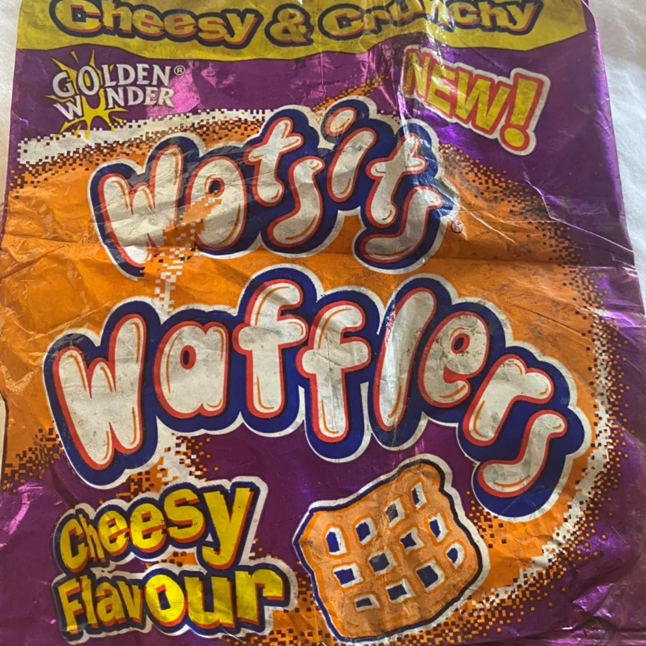 Close-up of a crumpled purple, orange and yellow snack packet printed with “Golden Wonder,” “Wotsits Wafflers,” “Cheesy Flavour,” “Cheesy & Crunchy” and “NEW!”, plus an illustration of a square lattice-shaped snack.