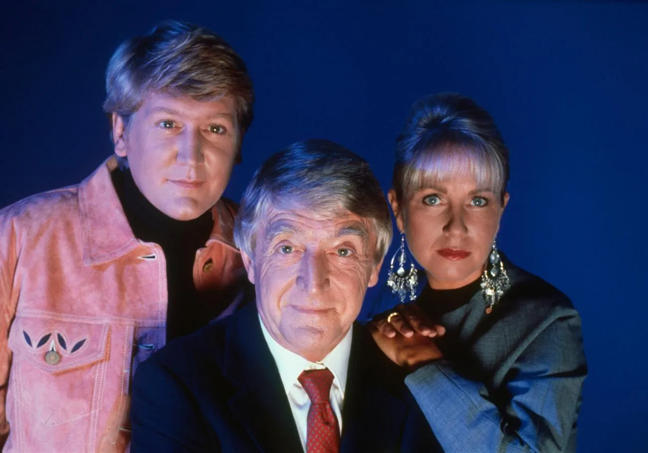 Vertical studio portrait of three people against a dark blue background: a grey-haired man in a dark suit and red tie sits in front, flanked by a man in a pink jacket and a blonde woman in a grey jacket with large earrings.
