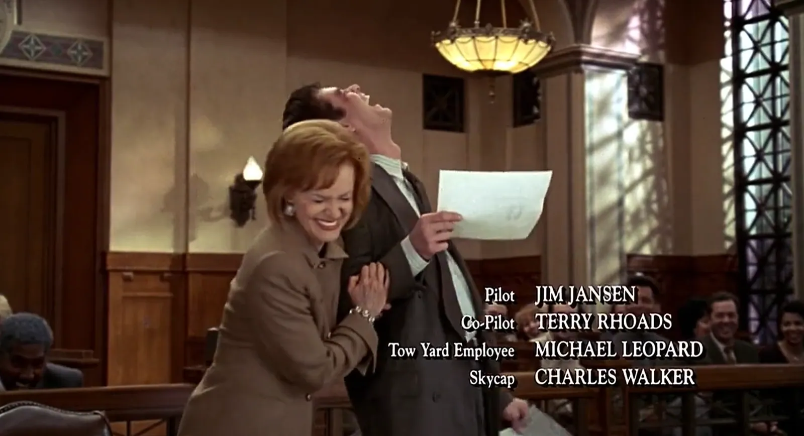 A red-haired woman and a dark-haired man laugh in a wood-panelled courtroom while the man holds a sheet of paper; white crew credits are superimposed across the lower-right portion of the scene.