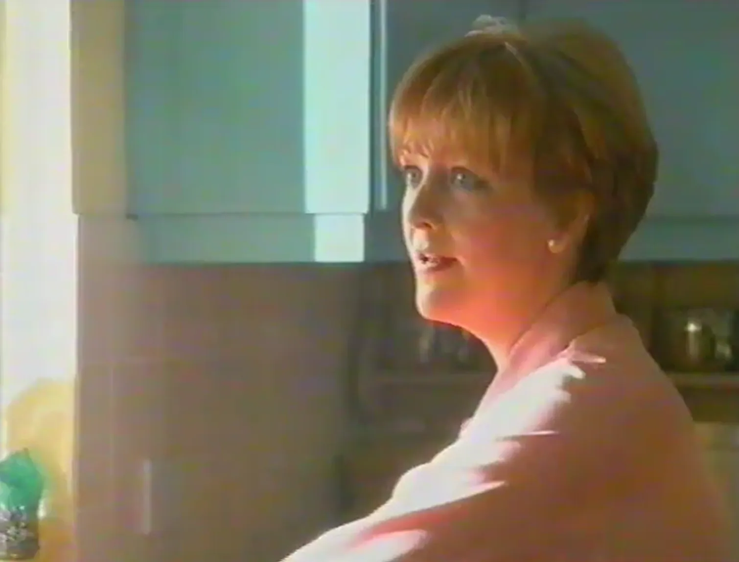 A short-haired woman wearing a pale pink top and a pearl stud earring looks to the left while standing in a kitchen with light-colored cabinets; bright light enters from the left edge.