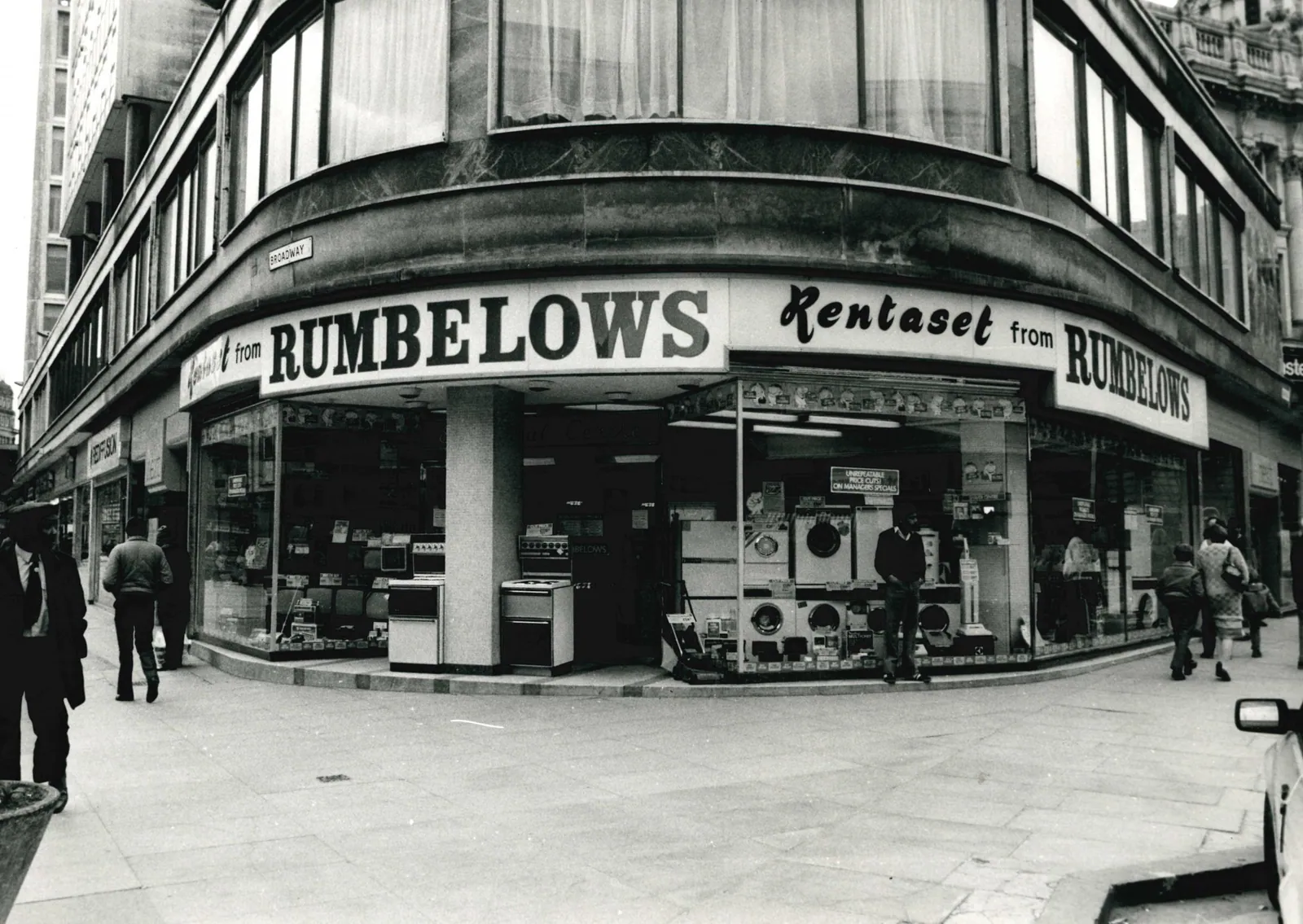 Black-and-white photograph of a corner Rumbelows electrical shop beneath a curved sign reading “RUMBELOWS” and “Rentaset from Rumbelows.” Televisions, cookers, washing machines and other appliances fill the display windows while pedestrians pass on the pavement; a street sign reads “Broadway.”