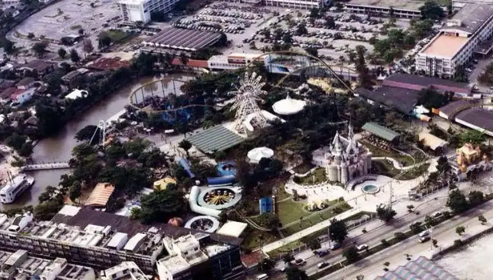 Elevated aerial view of Dan Neramit amusement park in Bangkok, showing landscaped grounds, amusement rides, winding pathways and surrounding roads and buildings.