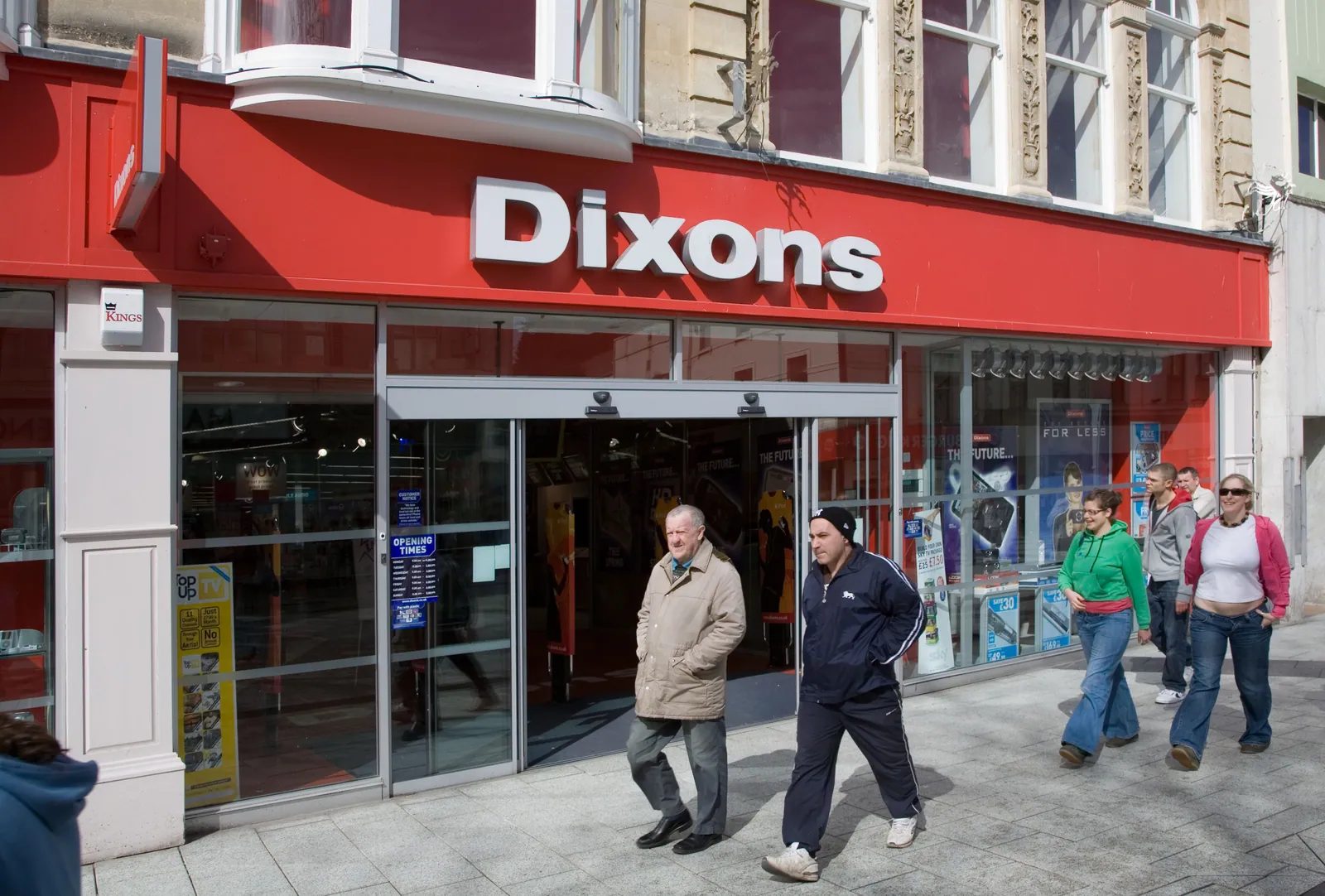 Pedestrians pass a Dixons shop in Cardiff, Wales, with a red fascia, large white lettering and electronics displayed behind the windows.