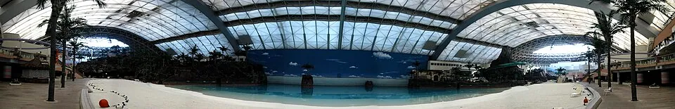 A wide panorama of an indoor beach and pool beneath arched roof beams, with palm trees at both sides.