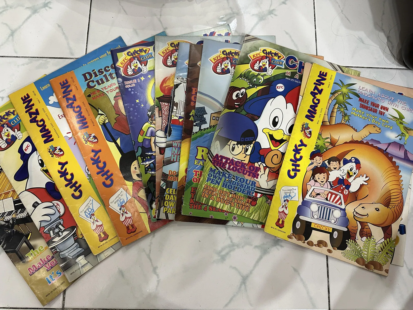 Several colourful KFC Chicky Club children's magazines arranged in a fan on a pale tiled floor, with illustrated covers featuring Chicky and other cartoon characters.