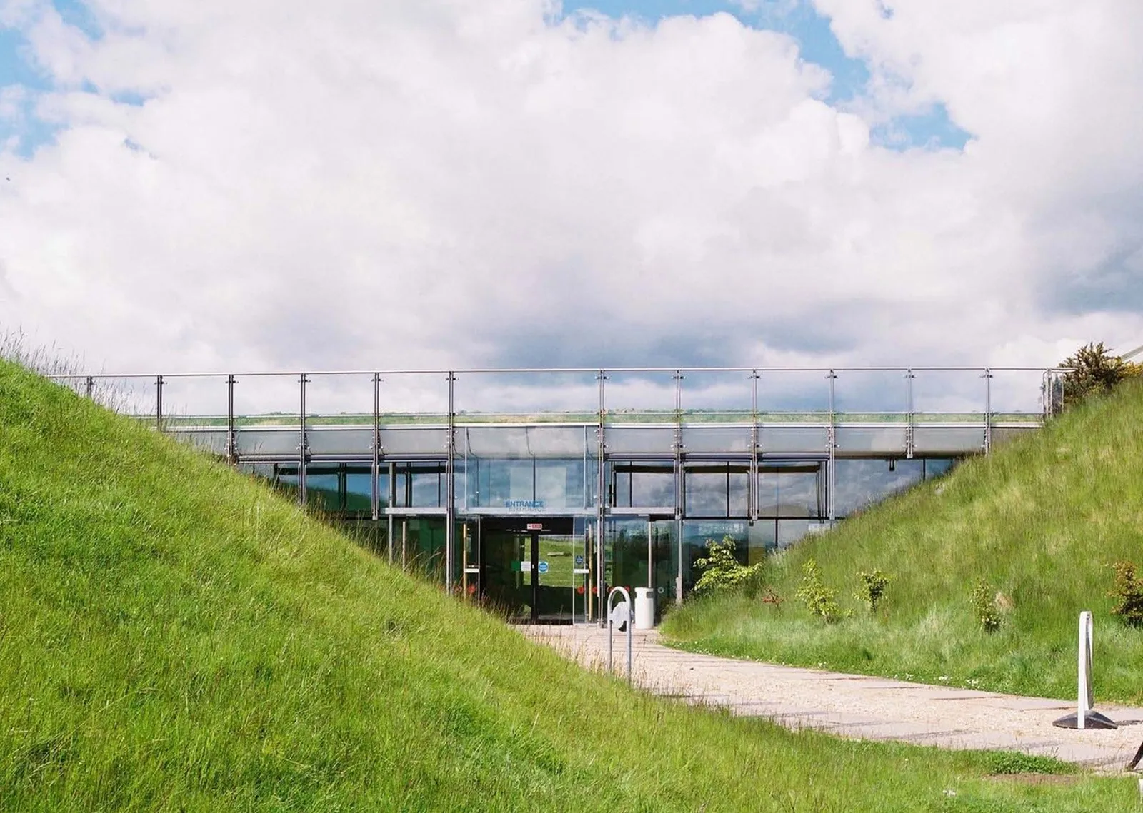 Glazed entrance beneath a metal-railed terrace, approached by a paved path between steep grass-covered banks under a cloudy sky.