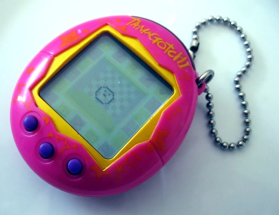 Pink and yellow egg-shaped Tamagotchi with three purple buttons and a metal bead-chain, photographed on a pale background.