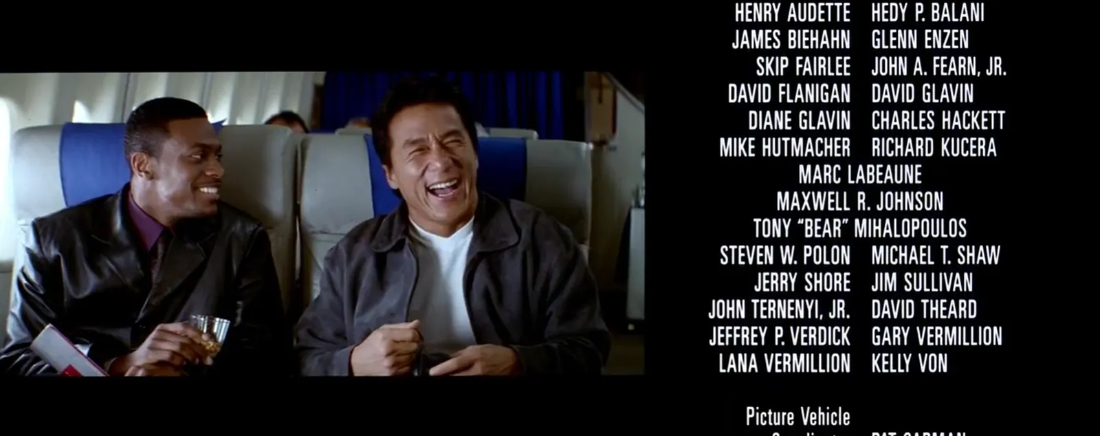 Two men seated beside each other in an aeroplane cabin smile and laugh in a video panel on the left, while columns of white production credits appear on black at right.