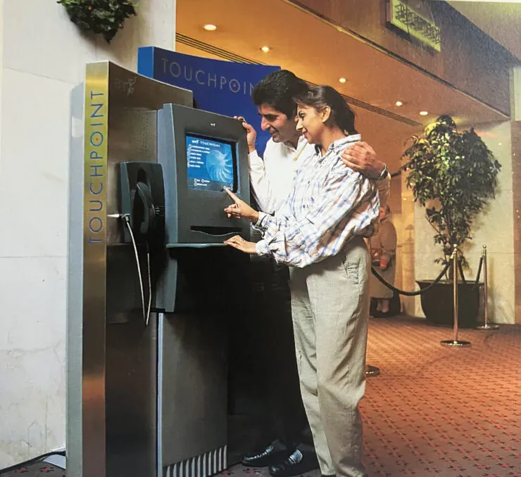 A woman operates a tall BT Touchpoint kiosk while a man stands beside her with one arm around her shoulders. The kiosk has a small color screen, a keyboard-like ledge, a telephone handset on its left side, and vertical TOUCHPOINT branding.