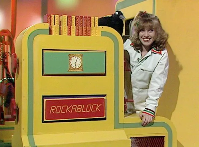 Television studio still of a smiling woman in a white outfit standing behind a large yellow and green structure with upright patterned blocks, a small clock, and a red panel reading “ROCKABLOCK”; a black bird figure is behind her.