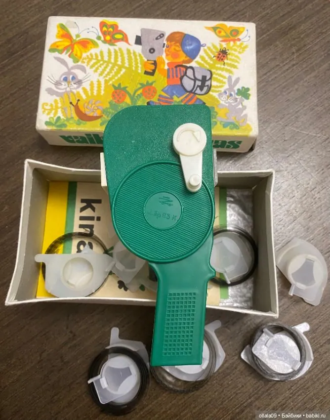Green hand-cranked Neringa film viewer arranged with its illustrated box, leaflet and circular film holders.