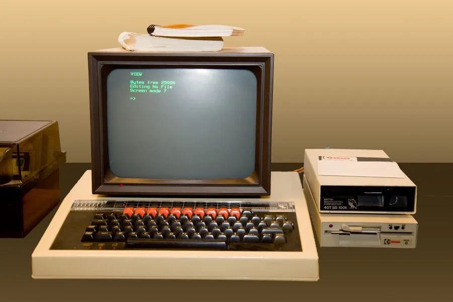 Colour photograph of a beige BBC Micro beneath a CRT monitor displaying green text, with a book resting on the monitor and two external disk drives positioned to the right.